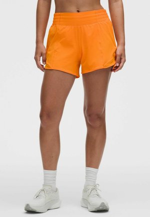 Fit person wearing bright orange athletic shorts, white crew socks, and white running shoes, standing against a plain light background.