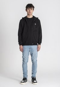 Sudadera negra con cremallera y bolsillo frontal, usada sobre una camiseta negra, combinada con jeans de mezclilla azul claro y zapatillas negras y blancas.