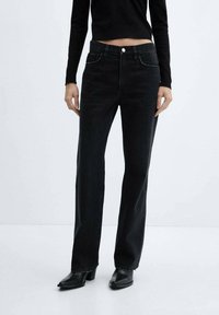 Jeans de mezclilla negra con diseño de pierna recta, bolsillos frontales y cierre de botón visible. Llevados con una camiseta ajustada negra y botas de tacón.