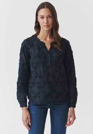Blusa blu navy con maniche lunghe, ricamo floreale testurizzato, scollatura a V e tre bottoni sul davanti. Abbinata a jeans slim in denim.