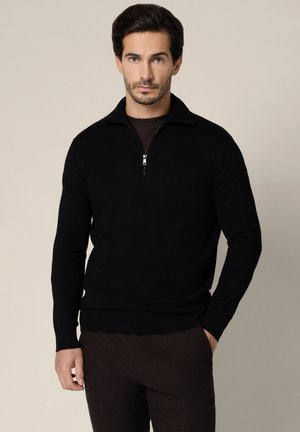 Mężczyzna o ciemnych włosach i brodzie, ubrany w czarny sweter z zamkiem do połowy długości i ciemne spodnie, stoi na tle beżowego tła, z jedną ręką w kieszeni.