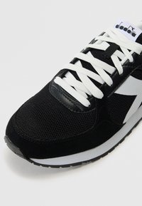 Schwarze Sportsneaker aus Mesh und Wildleder, mit weißen Schnürsenkeln und einer grauen Sohle sowie einem auffälligen weißen Logo an der Seite.