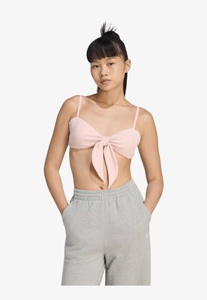 Giovane donna che indossa un crop top rosa chiaro annodato e pantaloni della tuta grigi con le mani nelle tasche, in piedi davanti a uno sfondo bianco semplice.
