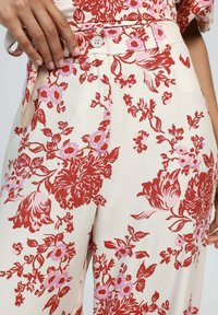 Pantalons à imprimé floral en crème avec des motifs rouges et roses. Présente une fermeture à bouton et une texture de tissu douce.