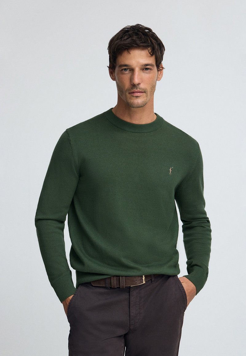 Waldgrüner Pullover mit Rundhalsausschnitt, gerippten Bündchen und subtiler Logodetail auf der Brust, kombiniert mit dunkelbraunen Hosen und einem Gürtel.
