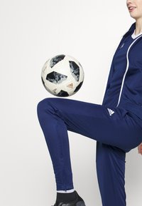 Námornícka súprava Adidas s bielymi akcentmi; priliehajúce nohavice s oproti zipsom. Na kolene je vyvážený vzorovaný čierno-biely futbalový lob.