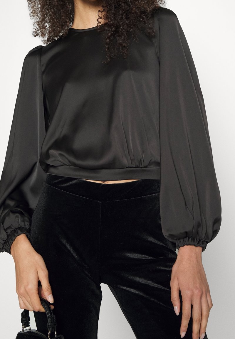 Donna che indossa una blusa nera di satin a maniche lunghe con maniche a sbuffo e pantaloni di velluto neri, mentre tiene una piccola borsa nera.
