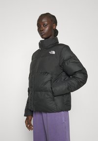 Giacca a vento nera con colletto alto, caratterizzata da baffle cuciti e una cerniera frontale. Il logo The North Face è visibile sul petto sinistro.