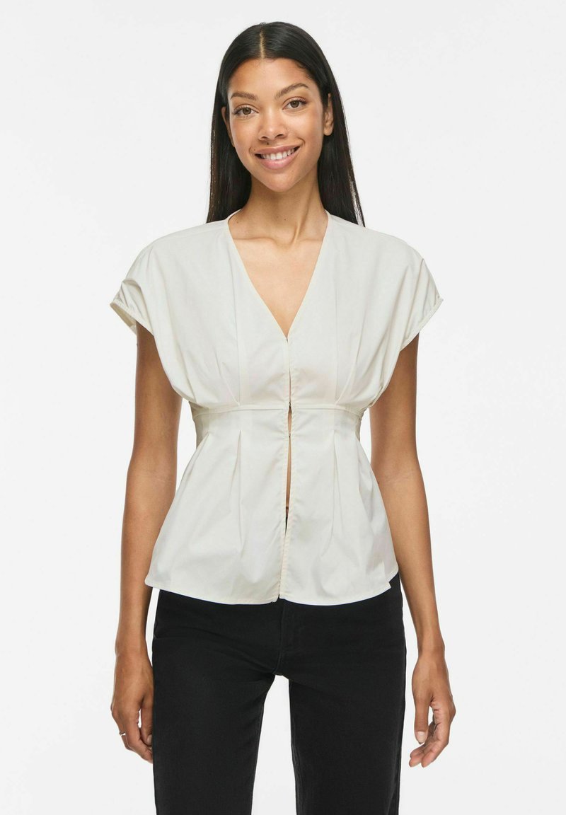 Femme souriante aux longs cheveux foncés, portant un chemisier blanc à manches courtes et col en V, ainsi qu'un pantalon noir, sur un fond blanc uni.