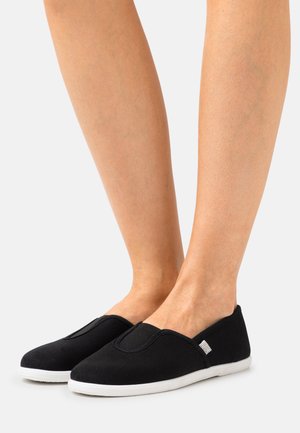 Sapatos slip-on pretos em tela com painéis laterais elásticos e solas de borracha brancas; textura lisa, forma arredondada, detalhes mínimos.
