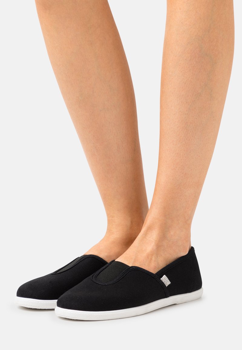 Sapatos slip-on pretos em tela com painéis laterais elásticos e solas de borracha brancas; textura lisa, forma arredondada, detalhes mínimos.