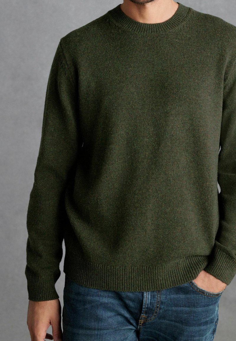 Homme portant un pull en maille vert foncé et un jean bleu, une main dans la poche, sur fond gris.