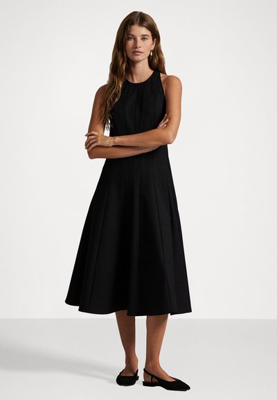 Ženska stoji z crossed arms, oblečena v brezrokavni črni midi obleki in črnih slingback balerinkah na enobarvni podlagi.