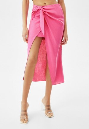 Vrouw draagt een roze wikkelrok met hoge split, gestrikt in de taille, en hoge hakken met transparante bandjes, staand tegen een witte achtergrond.