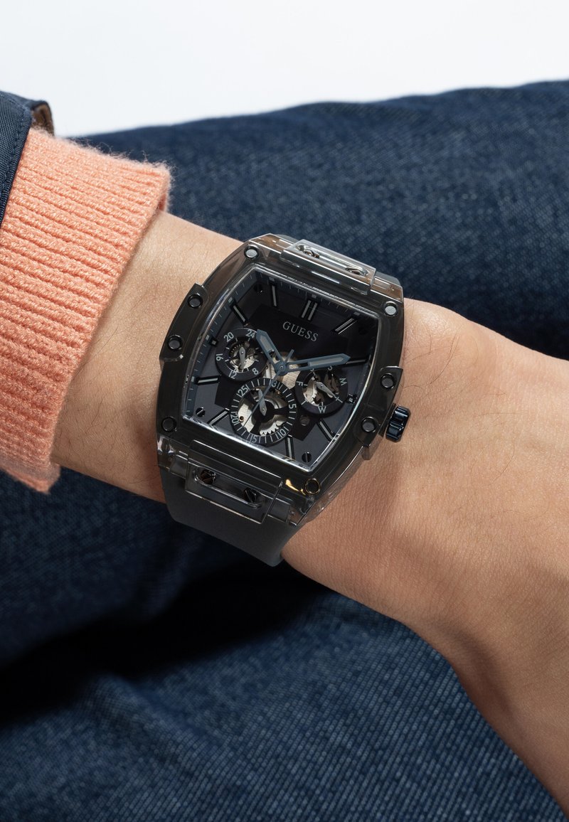 Zapešće na kojem je nošen crni Guess sat sa kvadratnim brojčanikom, analognim prikazom, više brojčanika i srebrnim detaljima, upareno s breskvastim džemperom i tamnim trapericama.