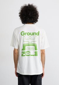 Hvid bomulds t-shirt med en stor grøn grafisk tryk på ryggen, der har teksten "Ground YOUR SOLE" og et stiliseret figurbillede.