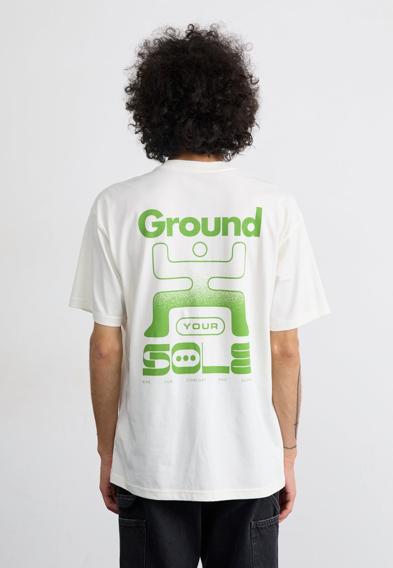 Hvid bomulds t-shirt med en stor grøn grafisk tryk på ryggen, der har teksten "Ground YOUR SOLE" og et stiliseret figurbillede.