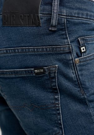 Tumši zili džinsi ar teksturētu apdari. Izceltas ar melnu ādas birku ar uzrakstu "BIG STAR" un mazu logotipa tagu aizmugures kabatā.