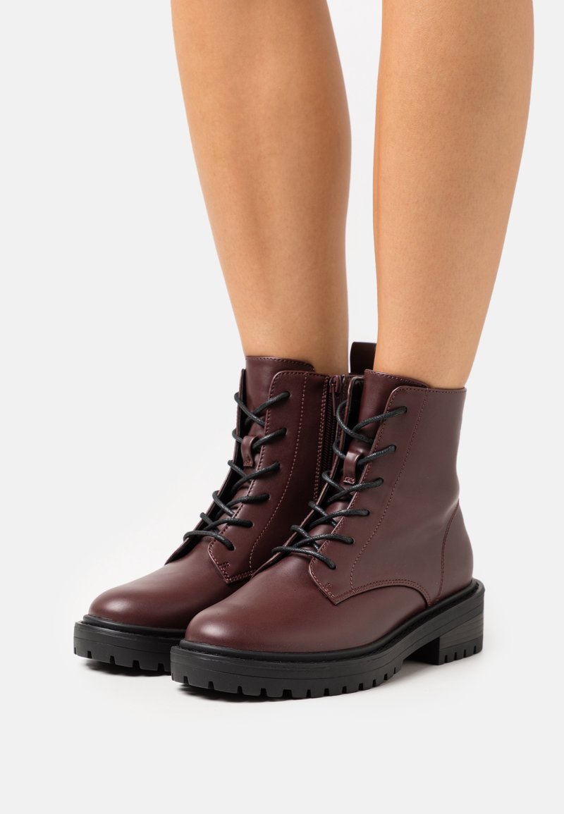 Jambes portant des bottines en cuir marron foncé à lacets, semelles épaisses en caoutchouc noir et zips latéraux sur fond blanc.