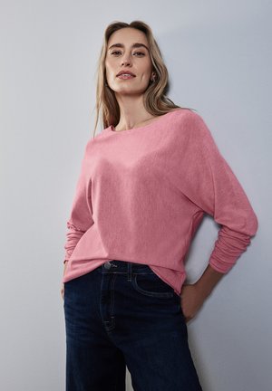 Pull rose clair à manches longues avec une coupe décontractée, texture douce et décolleté large, associé à un jean bleu foncé.