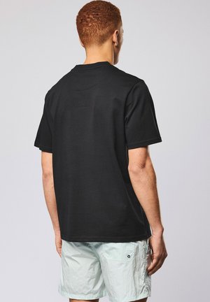 Mann mit kurzem lockigem Haar, der ein einfaches schwarzes T-Shirt mit kurzen Ärmeln und hellfarbene Shorts trägt, von hinten vor einem grauen Hintergrund.