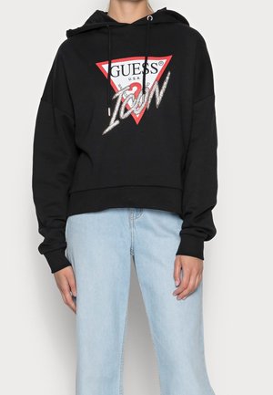 Osoba nosząca czarną bluzę z kapturem z czerwono-białym logo Guess i napisem "Icon", w połączeniu z jasnoniebieskimi dżinsami.