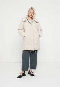 Casaco acolchoado bege com gola alta, fechos de botão e bolsos laterais. Usado sobre calças largas cinza e sapatos planos pretos.