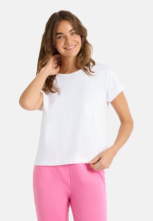 Junge Frau mit welligem braunem Haar, die ein weißes Kurzarmhemd und rosa Hosen trägt, lächelnd mit einer Hand am Hals.