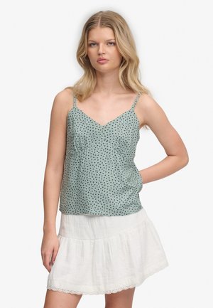 Jeune femme aux cheveux blonds portant un haut vert à pois sans manches et une jupe blanche à volants, debout avec une main sur la hanche.