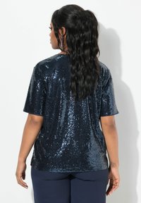 Top de lentejuelas azul marino con mangas cortas, ajuste relajado y superficie texturizada, mostrado desde la parte posterior sobre un fondo blanco.