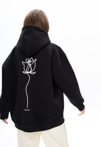 Zwarte oversized hoodie met een grote witte rozenborduursel op de achterkant, met lange mouwen en een capuchon met trekkoord.