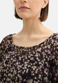 Durchsichtige schwarze Bluse mit rundem Ausschnitt, versehen mit einem mehrfarbigen, abstrakten Punktmuster in Lila, Beige und Braun. Strukturiertes Gewebe.