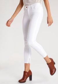 Jeans skinny de denim blanco con un diseño ajustado, que presentan un detalle de cremallera en el tobillo y un botón de cobre. Combinados con botas de tacón marrones hasta el tobillo.