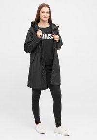 Zwarte waterproof regenjas met capuchon, drukknoopsluitingen en een relaxte pasvorm, gedragen over een zwarte sweatshirt en strakke zwarte broek. Witte sneakers.