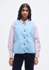 Ανοιχτό μπλε γιλέκο από φλις με στρογγυλεμένο λαιμόκοψη, κουμπιά μπροστά και δύο πλαϊνές τσέπες, συνδυασμένο με ριγέ ροζ πουκάμισο από κάτω.