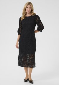 Black lace dress with puffed sleeves and a knee-length hem. Features floral patterns with a semi-sheer lower section. Styled with black shoes.