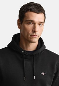 Sort sweatshirt med justerbar hætte, der har et GANT-logo. Stoffet virker blødt med en glat tekstur og en afslappet pasform.