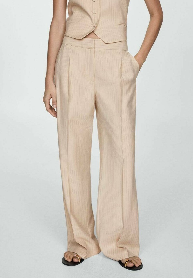 Pantalon beige à fines rayures, taille haute, poches latérales, coupe large, confectionné en tissu lisse. Associé à des sandales noires simples.