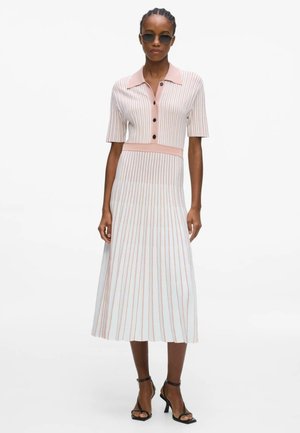 Kobieta ubrana w różowo-białą midi sukienkę w pionowe paski z guzikami i kołnierzykiem, czarne sandały na obcasie i okulary przeciwsłoneczne w okrągłym kształcie.