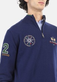 Navy blue zip-up sweater made of soft material, featuring embroidered logos, green accents, and a large white number "2" on the sleeve.