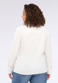 Camicia bianca a maniche lunghe con una texture liscia e un sottile dettaglio della spalla posteriore, abbinata a jeans blu. La camicia ha una vestibilità comoda.