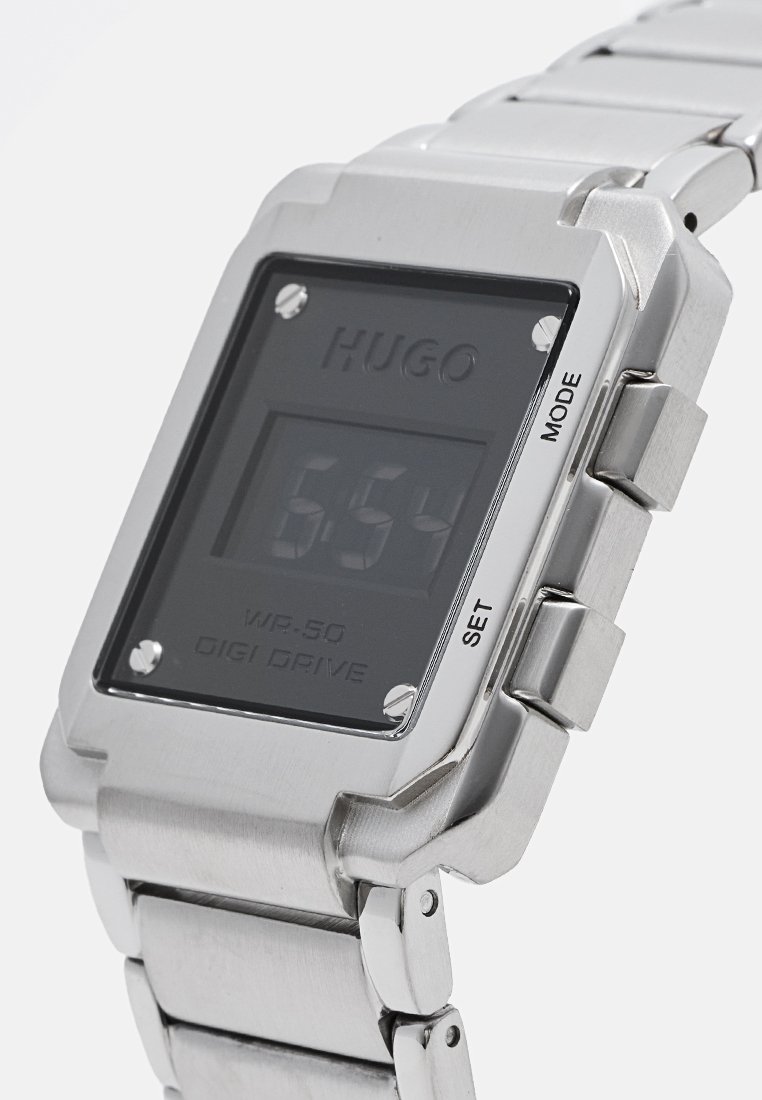 Reloj digital con pulsera de acero inoxidable, cara cuadrada negra, pantalla LCD y detalles en plata. Cuenta con botones etiquetados como 'AJUSTAR' y 'MODO'.