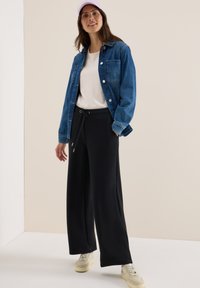 Veste en denim bleue, T-shirt blanc, pantalon de survêtement noir à jambes larges et baskets crème. Modèle portant une casquette rose clair, se tenant devant un fond neutre.