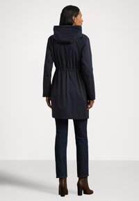 Dlouhý tmavě modrý kabát s kapucí a elastickým stahováním v pase, s dlouhými rukávy a volným střihem, doplněný tmavými džínami a kozačkami na podpatku.