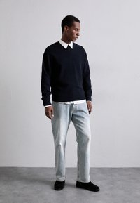 Marineblauer Rippstrickpullover mit Rundhalsausschnitt, über einem weißen Hemd mit Kragen getragen, kombiniert mit hellblauen Jeans und schwarzen Schuhen.