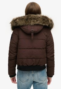 Bruine donzen jas met een capuchon met bontrand, geribbelde manchetten en zoom. Bevat doorgestikte delen en een detail met een rugband. Draagt bij lichte blauwe jeans.