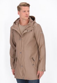 Svetlo hnedá nepremokavá bunda s kapucňou, zapínanie na patentky vpredu, dve bočné vrecká a hladká textúra, kombinovaná s béžovou košeľou a modrými džínsy.