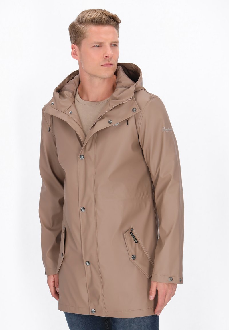 Svetlo hnedá nepremokavá bunda s kapucňou, zapínanie na patentky vpredu, dve bočné vrecká a hladká textúra, kombinovaná s béžovou košeľou a modrými džínsy.