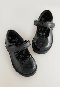 Chaussures en cuir verni noir avec un bout arrondi, présentant un design de chat sur le capuchon et une sangle en Velcro. Semelle intérieure noire à motifs.