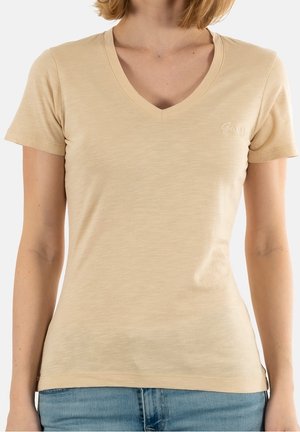 Kobieta ubrana w beżowy T-shirt z krótkim rękawem i dekoltem w serek oraz z małym logo "Guess" na piersi, zestawiona z niebieskimi dżinsami.